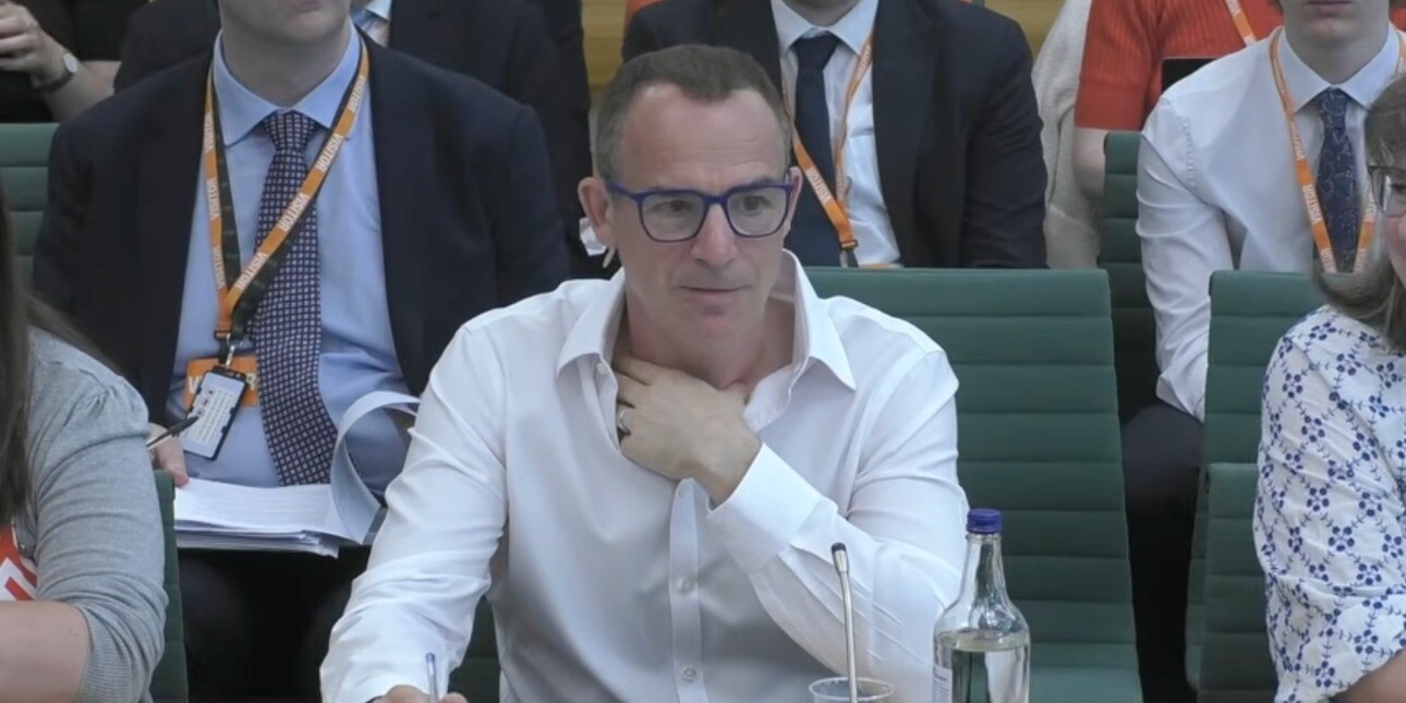 Martin Lewis sat in a green chair. Lewis is a white man with short dark hair, wearing glasses and a white shirt. He is surrounded by several people sat partially obscured either side of and behind him.