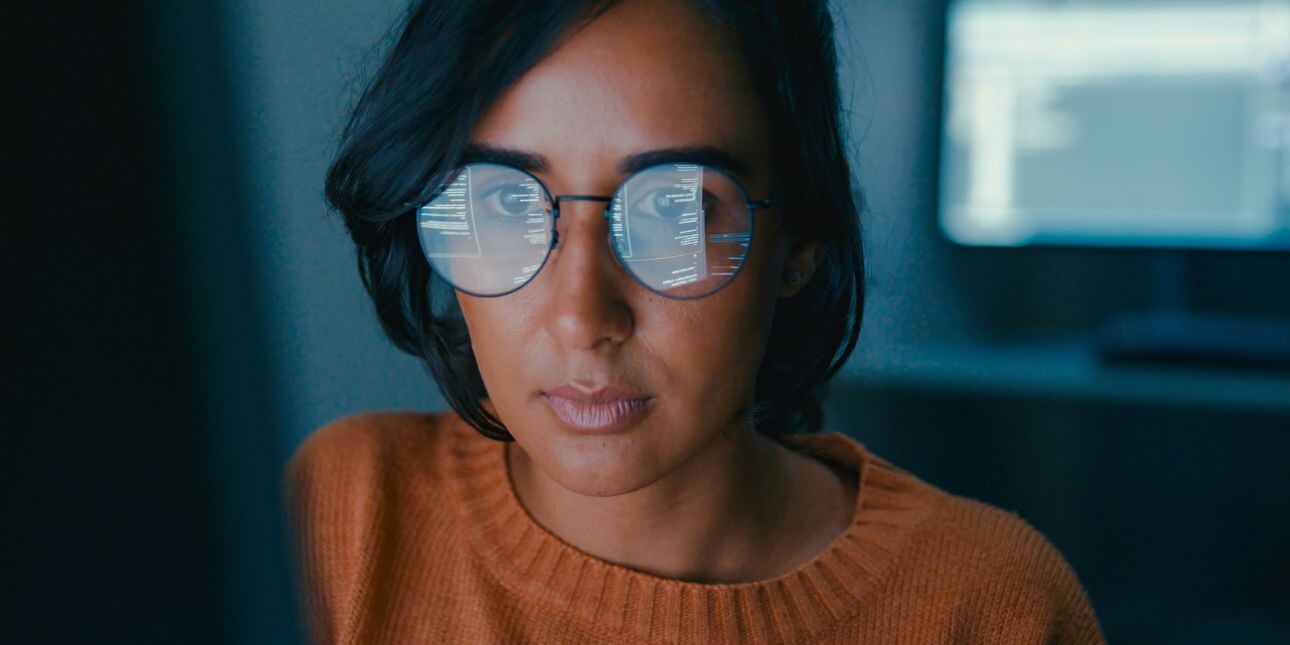 A woman with short dark hair and orange jumper is sat in front of her a computer screen which is reflected in her glasses.