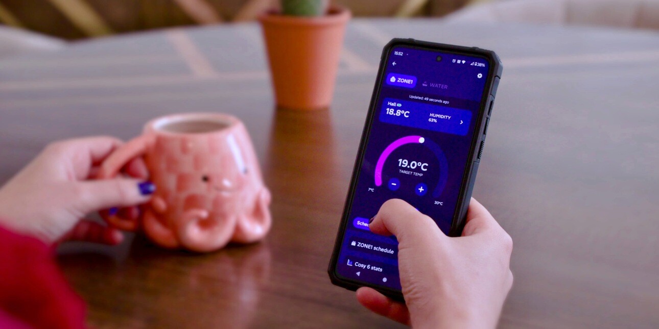A close up of a white hand holding a mobile phone which displays an energy app. To the left are a pink octopus shaped mug and a small house plant.