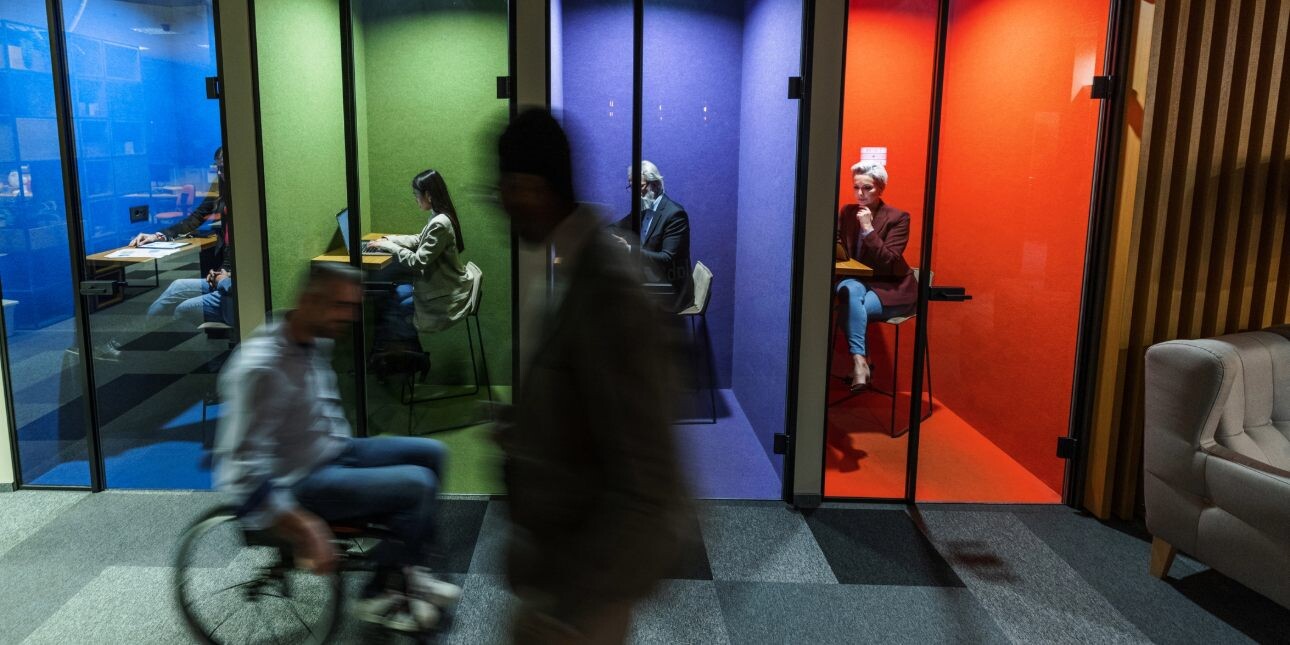 Four colleagues work individually inside multi-coloured booths while two silhouetted colleagues - one walking, one using a wheelchair - pass outside.