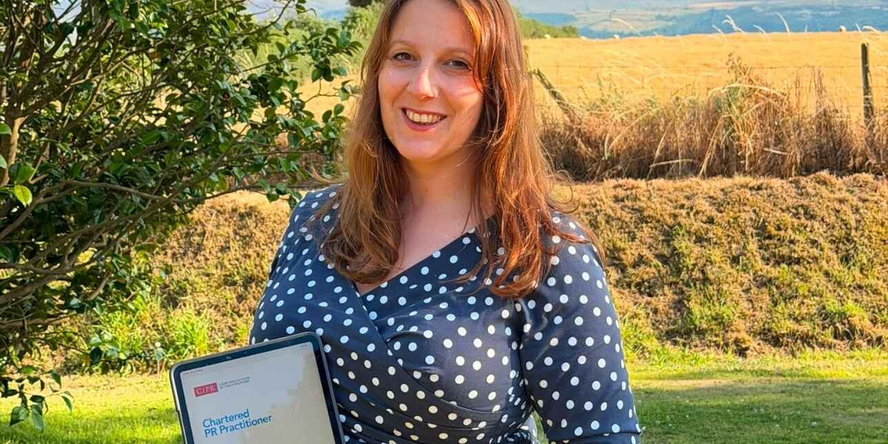 Kirstie Nickson - a white woman with long red hair, who is wearing a white spotted blue dress - holds a framed certificate in a sunny garden.