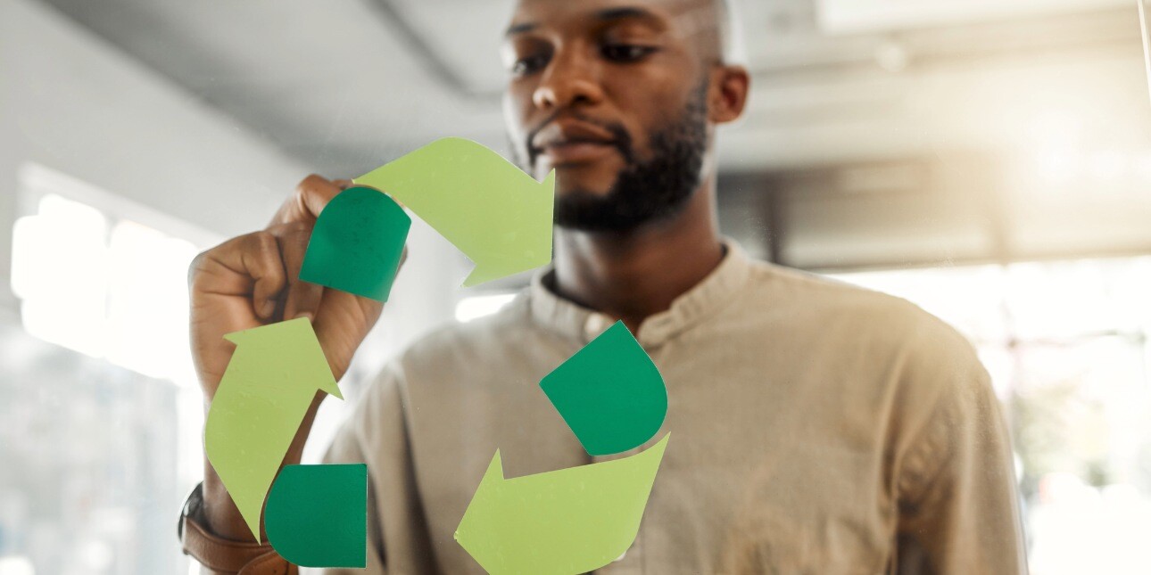 A Black man in an office, wearing a beige shirt, fixes a green recycling icon to a pane of glass.