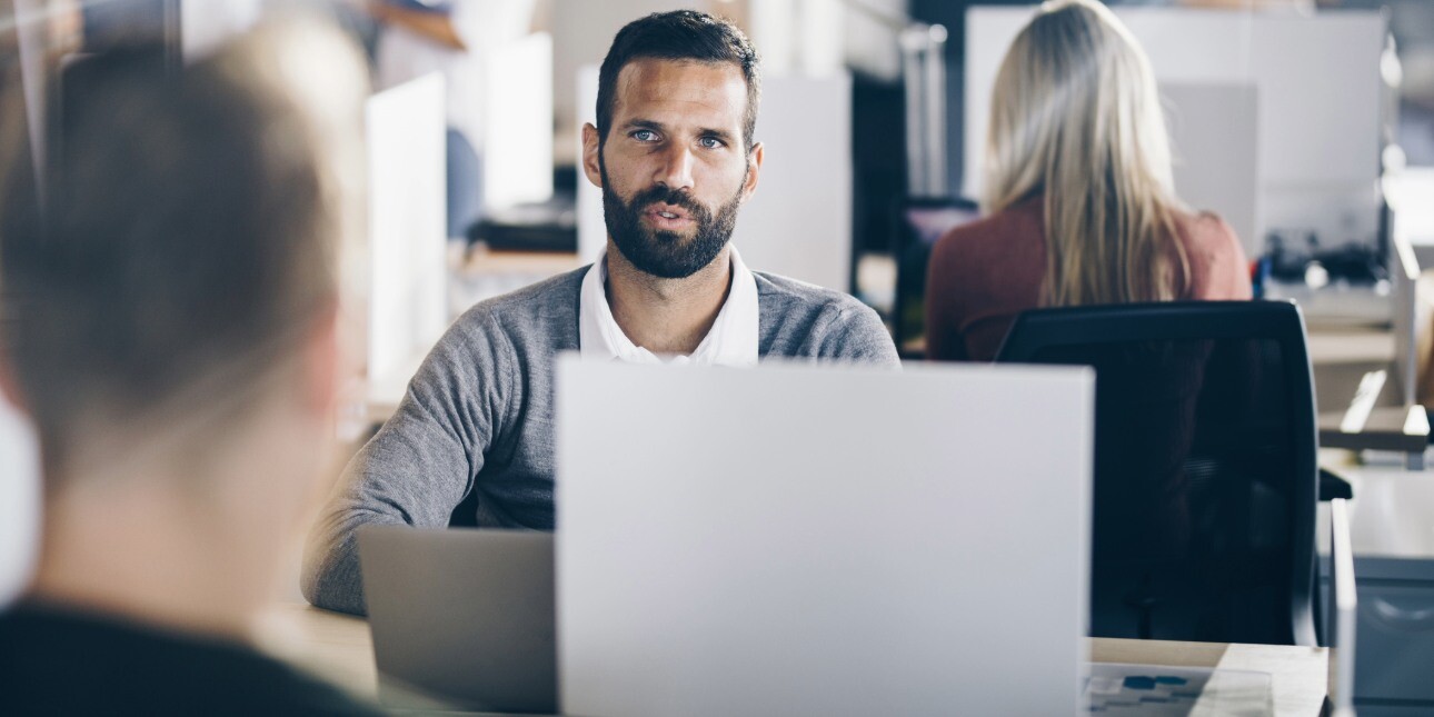 A white man with dark hair and beard sat at an office desk in conversation with a colleague whose back faces the camera.