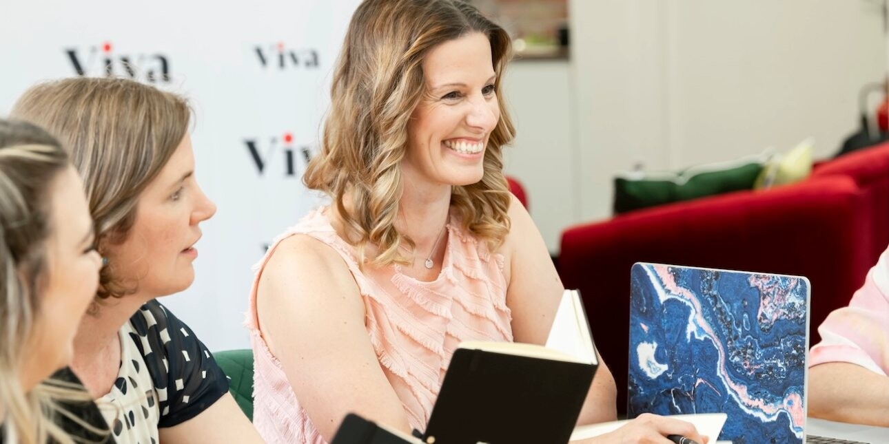 A smiling Lisa Egan-Woodhouse sat among members of the Viva team. Egan-Woodhouse is a white woman with shoulder length light brown hair, who is wearing a pink sleeveless top.