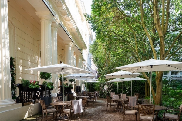 The terrace of 116 Pall Mall consisting of tables and chairs under parasols, leafy green trees on the right and three regency period columns of a cream building