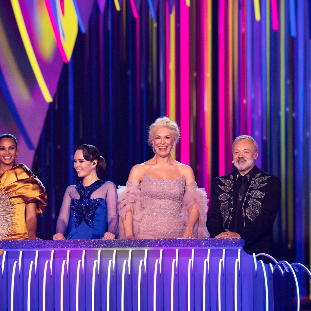 Alesha Dixon, Julia Sanina, Hannah Waddingham and Graham Norton on stage at the Eurovision Song Contest final. All are smiling and wear colourful clothing. The backgdrop is a mix of purple, yellow and pink