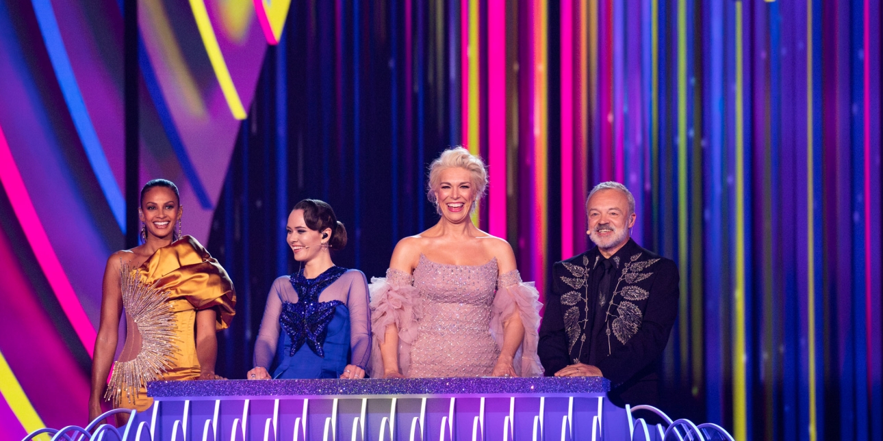 Alesha Dixon, Julia Sanina, Hannah Waddingham and Graham Norton on stage at the Eurovision Song Contest final. All are smiling and wear colourful clothing. The backgdrop is a mix of purple, yellow and pink