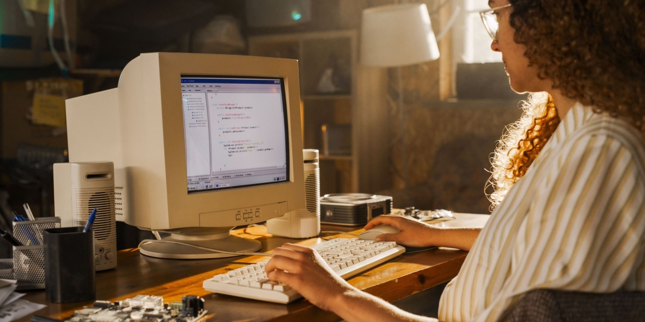 A women with brown curly hair sits in front of a large desktop computer monitor from the 1990s or early 2000s with separate speakers and a keyboard. The style of on screen graphics confirm this approximate date range