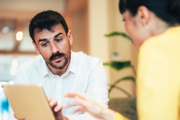 Two colleagues - a white man with dark hair and moustache - holds an iPad while in conversation with a female colleague with dark hair whose face is obscured.
