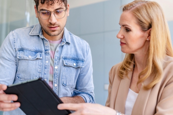 In a bright office, a young white man with dark curly hair and blue shirt holds a tablet while in conversation with a more senior female white colleague with blond hair and beige jacket