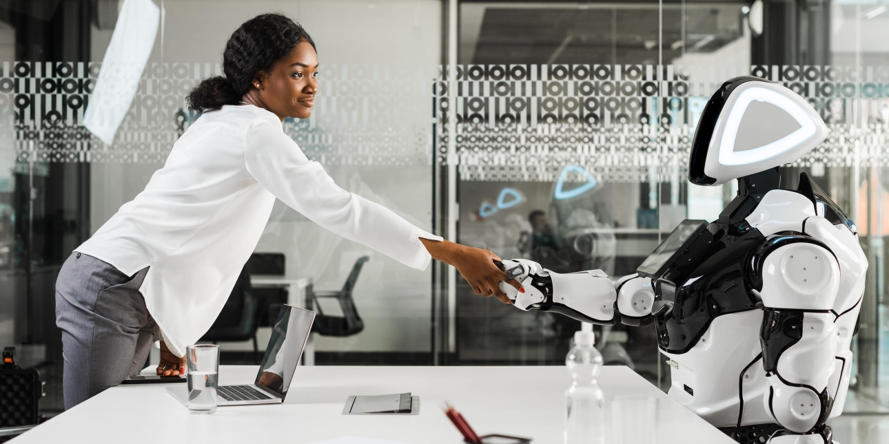 In a modern office with glass wall a Black business woman wearing a white shirt and grey trousers shakes with a white humanoid robot
