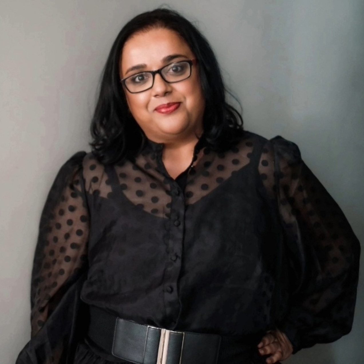 A colour portrait of Advita Patel against a grey backdrop. Advita is a British-Asian woman with shoulder length dark hair and glasses, who wears a black top.