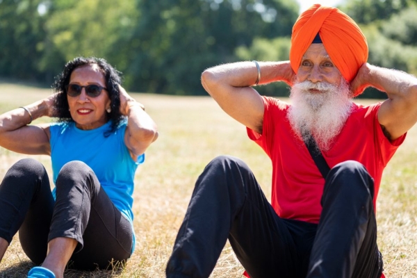 An elderly Asian couple perform sit ups on the grass. On the left is a woman wearing a blue vest and dark leggings. Next to her is a man wearing a red t-shirt, orange vest and blue tracksuit trousers.
