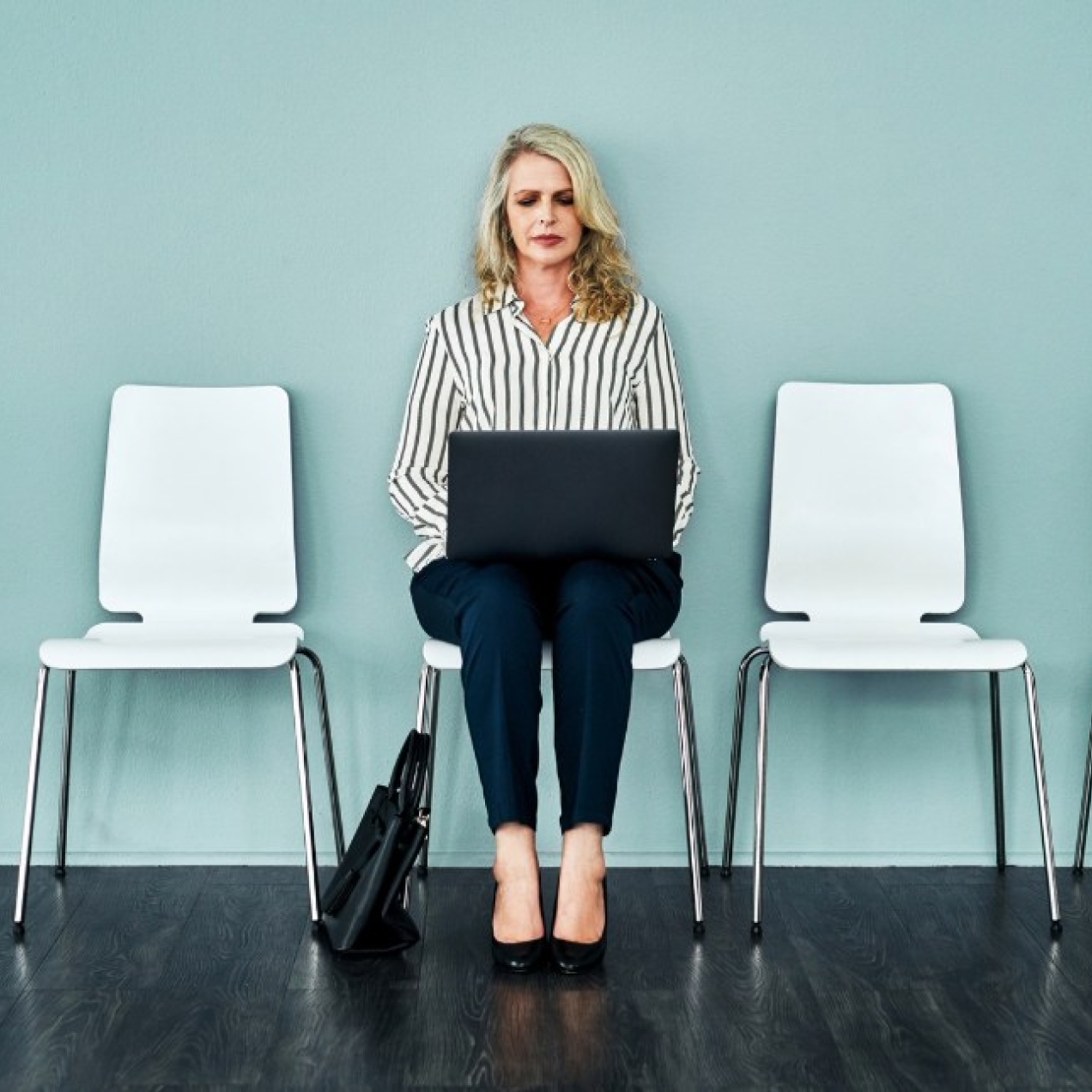 An older woman with blond hair and white top sits in a chair against a turquoise wall looking at the laptop resting on her knees. There are four empty chairs either side of her.