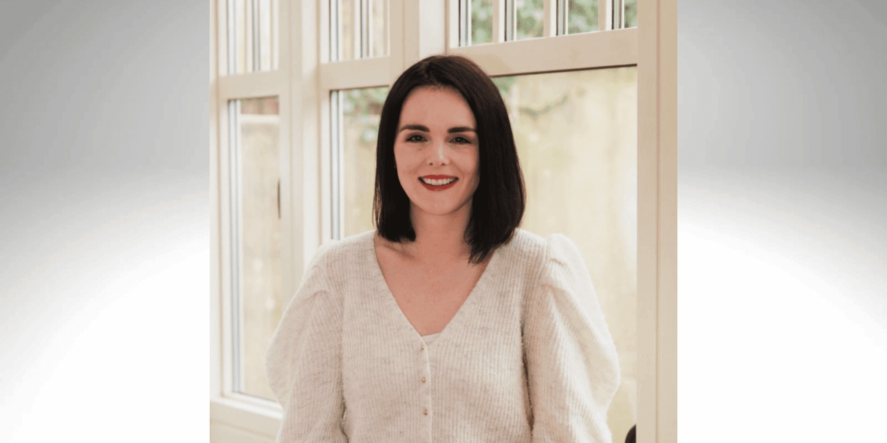 A portrait of a smiling Aishling O'Connor, a white woman with shoulder length dark hair and white top, stood in front of a window