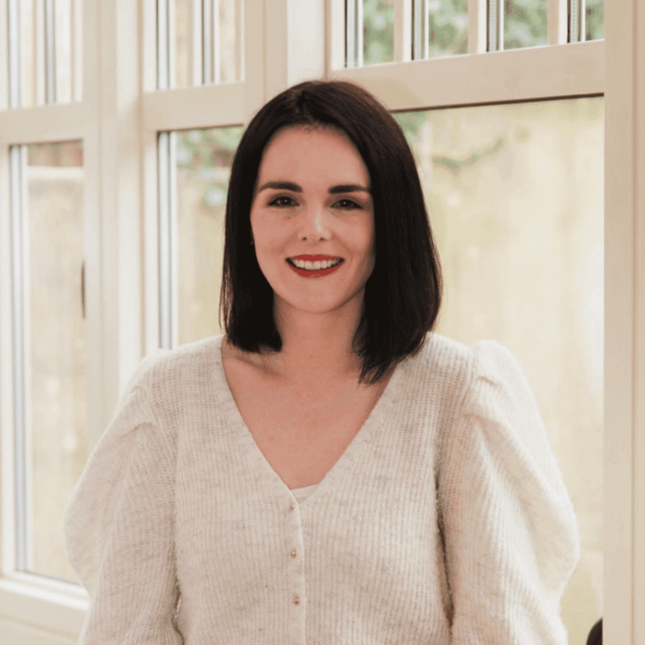 A portrait of a smiling Aishling O'Connor, a white woman with shoulder length dark hair and white top, stood in front of a window