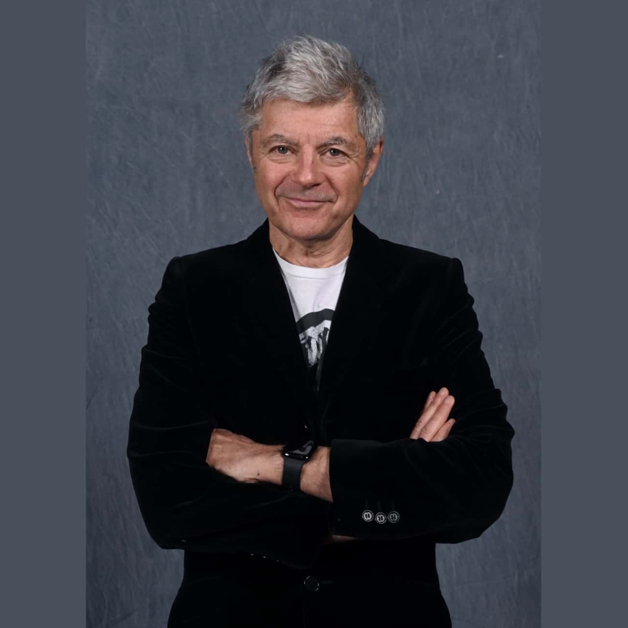 Alan Edwards, a white man with grey hair, wearing a black jacket over a white t-shirt. His arms are crossed. The background is grey