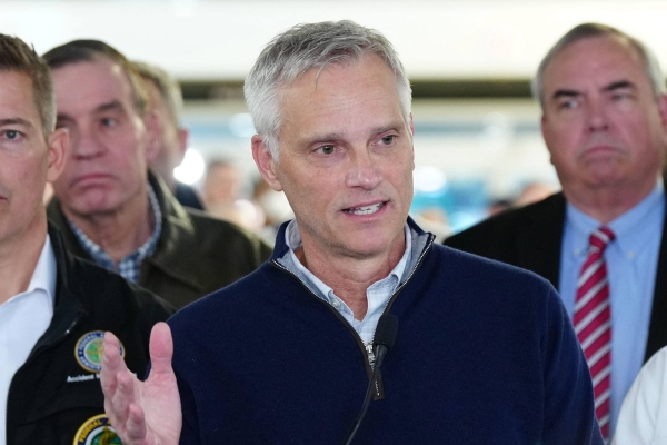 Robert Isom holding a press conference flanked by other officials. Isom is a white middle-aged man with short grey hair andwearing a navy zip-up sweater.