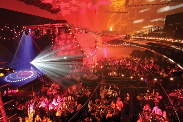 A colourful photo taken from a balcony overlooking the large auditorium and stages were audiences are sat around dozens of tables.