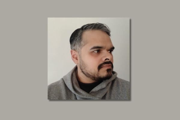 On a grey background, a colour side profile of Babar Khan Javed. Babar has short dark-grey hair and trimmed beard, and a grey hoody.