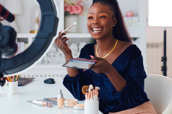A smiling beauty-influencer holding up a make-up brush at the camera which is mounted on a ring-flash. The influencer is a black woman wearing a blue top, sat at a table on which there is placed other make-up accessories