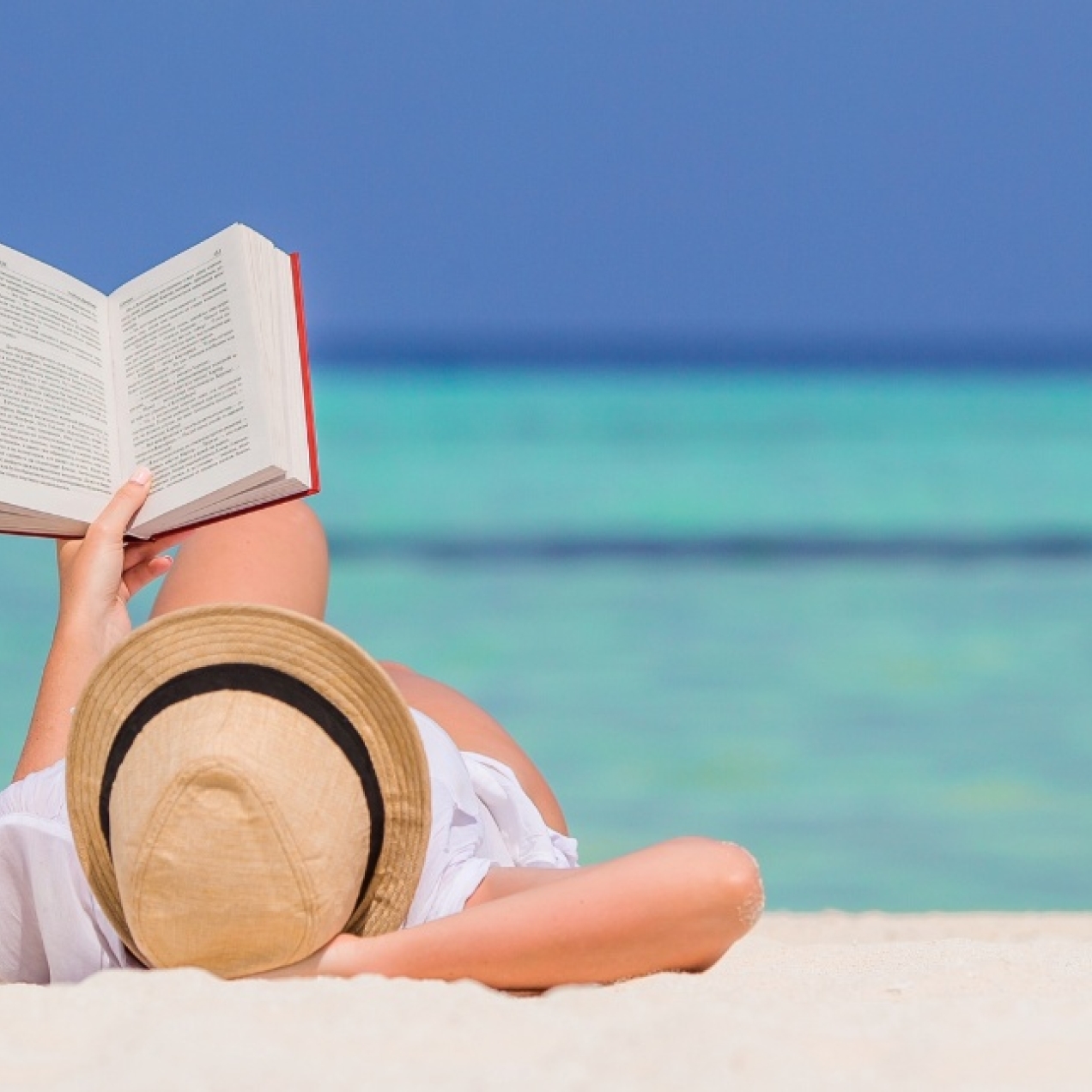 A low angle shot of a person wearing a hat lying on a sandy beach holding a book above them. Behind them are a two-tone blue sea and sky.