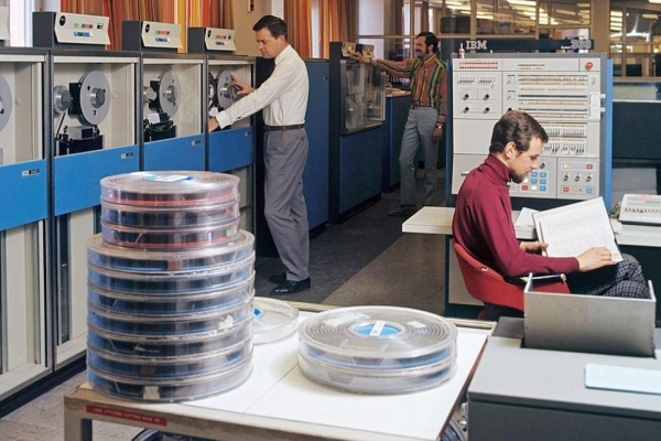 A photograph from 1966 showing three white men inside a room with early and large IBM computing equipment, stacks of data-tape reels and computer terminals.