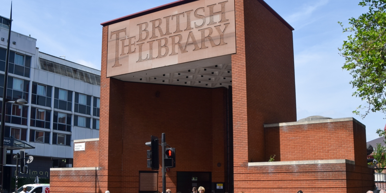 A tall red brick cuboid entrance with the words The British Library on the fascia of the lintel. The sky is blue and there are people and a pedestrian signal at the bottom of the image