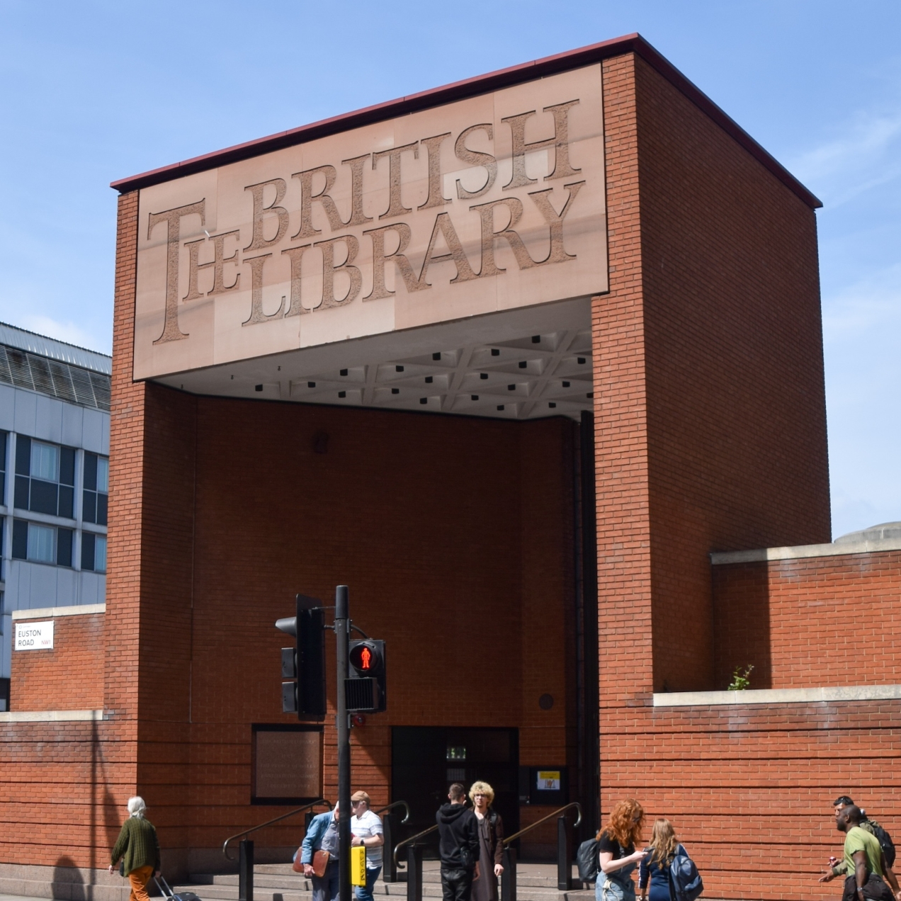 A tall red brick cuboid entrance with the words The British Library on the fascia of the lintel. The sky is blue and there are people and a pedestrian signal at the bottom of the image