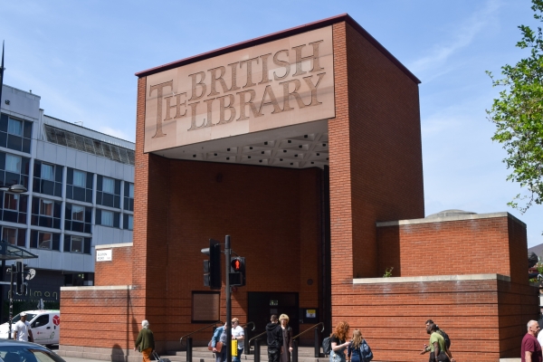 A tall red brick cuboid entrance with the words The British Library on the fascia of the lintel. The sky is blue and there are people and a pedestrian signal at the bottom of the image