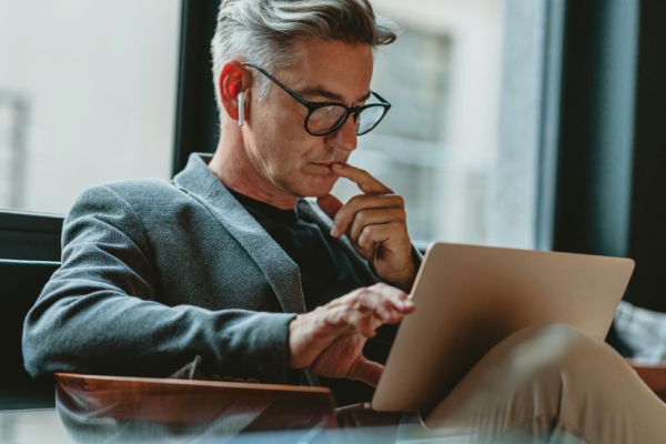 A white man with grey hair, wearing black-rimmed glasses, a grey jacket and a white headphone in his ear, stares at a laptop rested on his lap. He rests one finger on his chin and another on the laptop. There is a window in the background