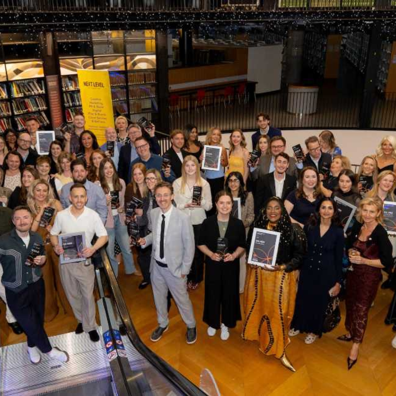 A photograph taken from the top of an escalator of dozens of smiling people holding framed certificates.