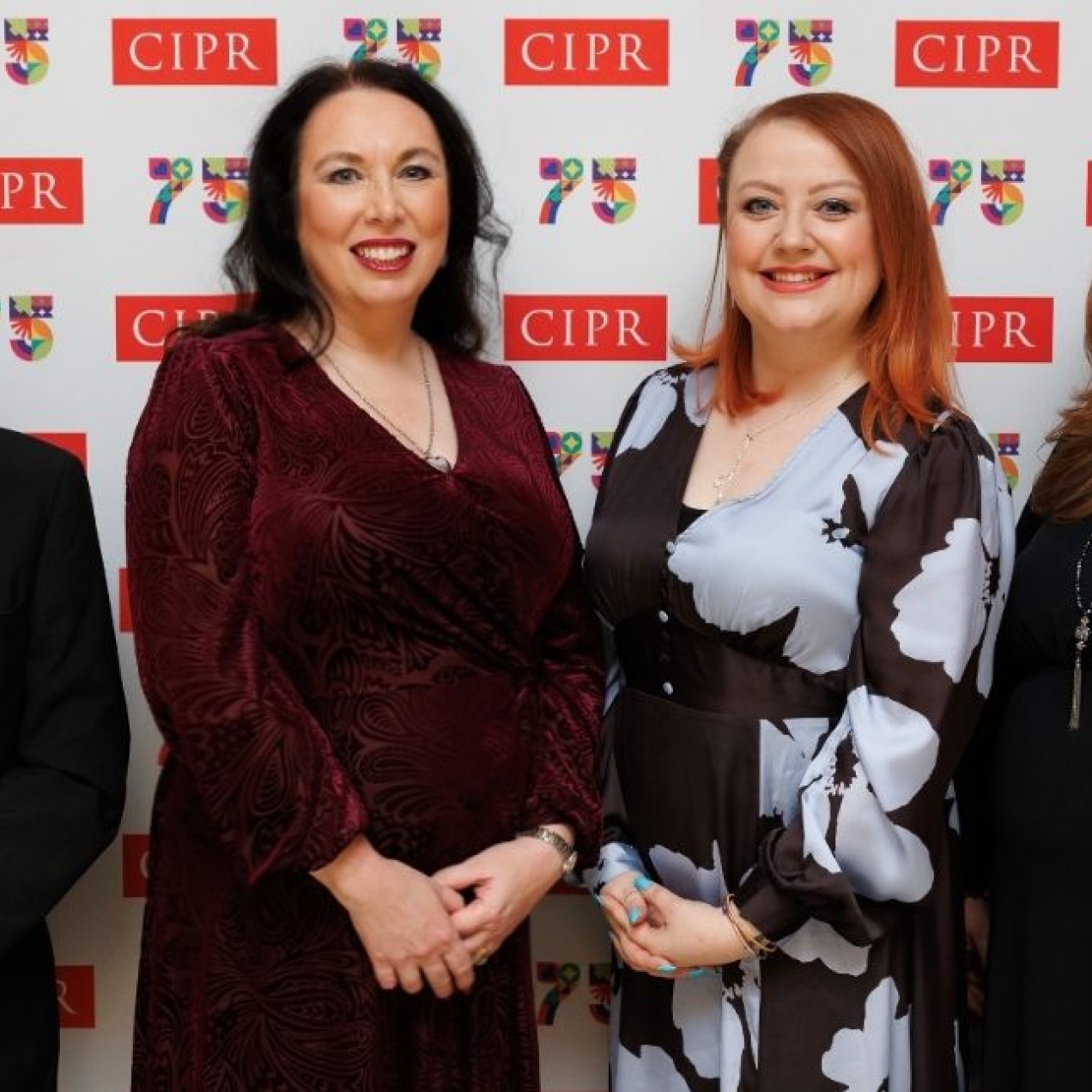 A white man and three white women wearing formal attire, stood smiling in front of a display board with the CIPR 75 logos.
