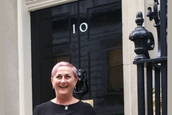 Carolyne Mitchell stood on the steps of 10 Downing Street. Carolyne is a white woman with short blonde hair wearing a black top. The door is back with white numbers