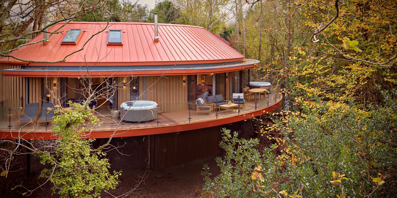 Exterior of a semi circular accommodation with red roof, wooden walls, balcony containing loungers and plunge pools, on stilts surrounded by tall leafy trees