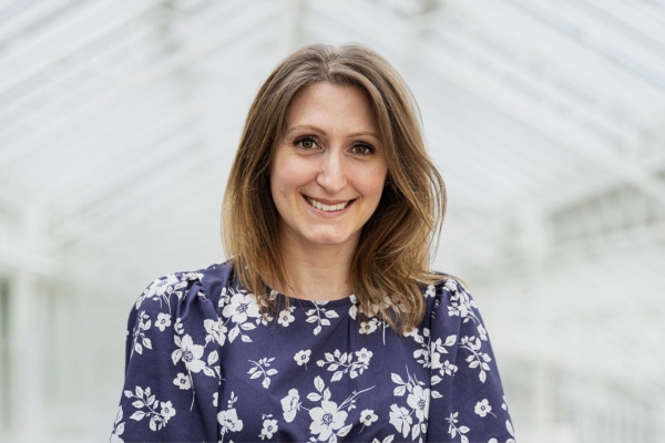 A portrait of Christine Richardson against the backdrop of a white pitched ceiling. Christine is a white woman with long dark hair who wears a blue top decorated with white florals