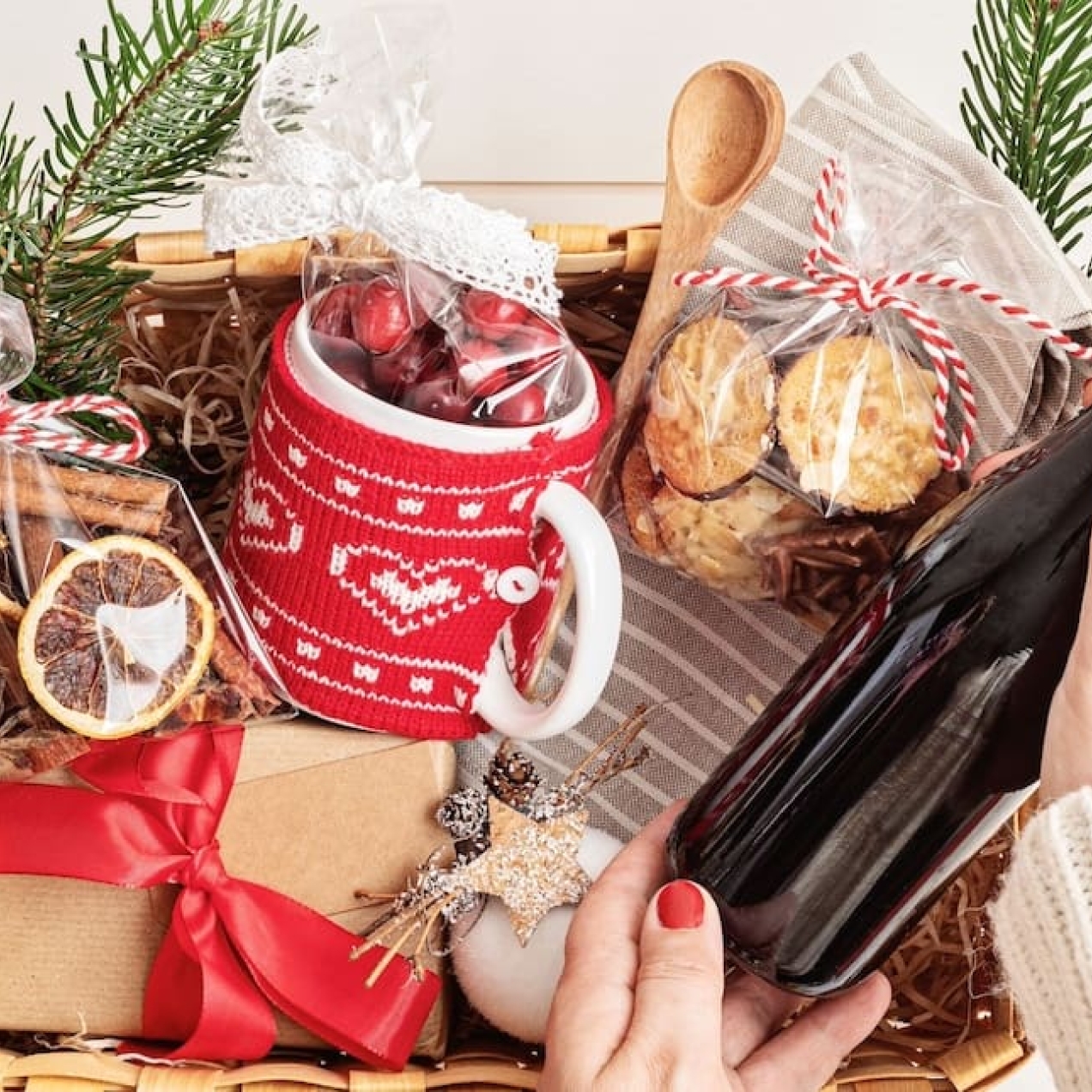 A Christmas hamper basket containing wrapped presents, ribbons, wooden spoons, biscuits and a mug covered in a red knitted cover. A white hand with red nail varnish lifts a bottle of red wine from the hamper