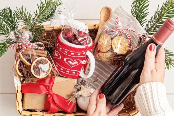 A Christmas hamper basket containing wrapped presents, ribbons, wooden spoons, biscuits and a mug covered in a red knitted cover. A white hand with red nail varnish lifts a bottle of red wine from the hamper