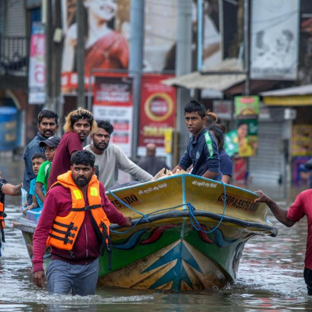 A group of rescuers wade through flooded streets as they pull a boat full of people they've rescued. There are buildings in the background.