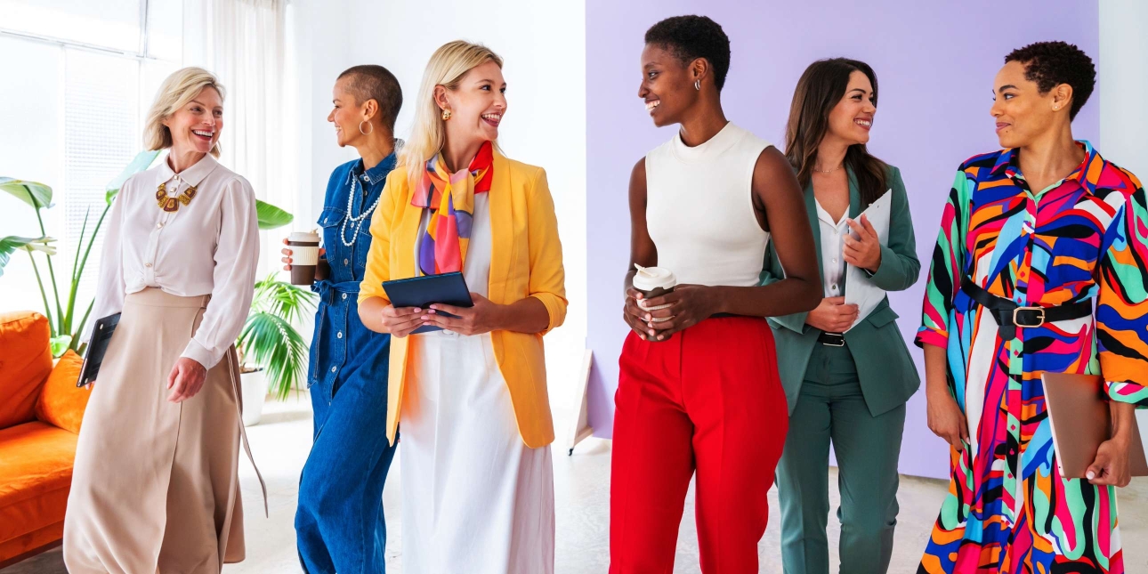Six smiling female colleagues of different ages and ethnicities walk in conversation with one another in a bright office. They are all wearing bright clothes.