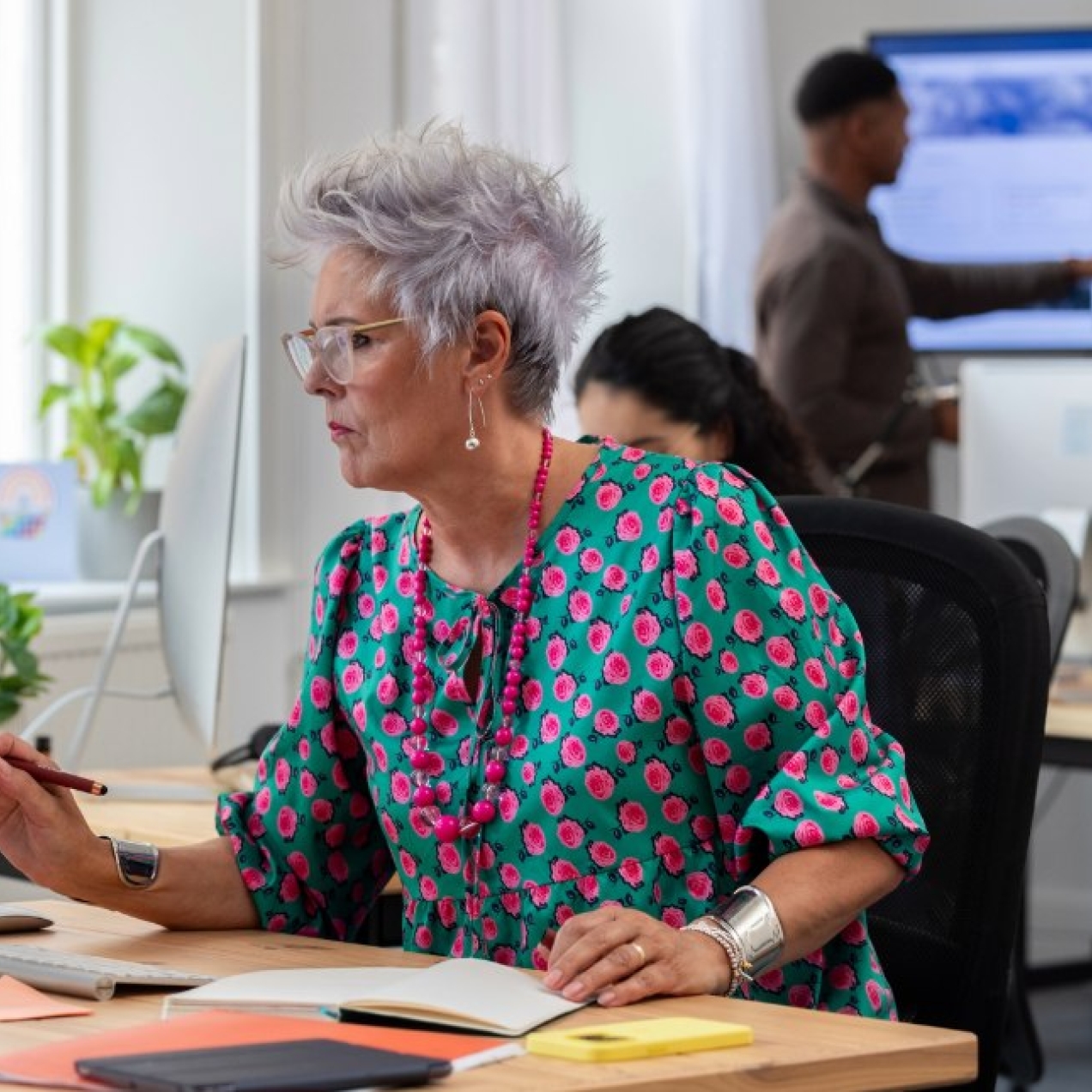 A colour photo of a middle-aged white woman with grey hair, glasses and a turquoise and pink dress sat at her desk in an office with paperwork and a monitor in front of her.