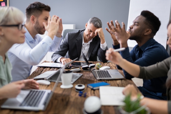 A suited white man leans his elbow on the table and rests his head while four colleagues of different genders, ages and ethnicities wave their arms in an angry discussion