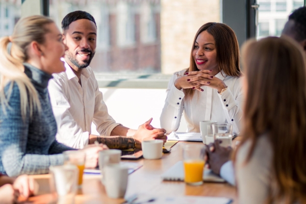 Six colleagues around board table. In focus are a white woman with blonde hair and blue shirt, a Black man with short dark hair and cream shirt, and a Black woman with brown hair and white shirt