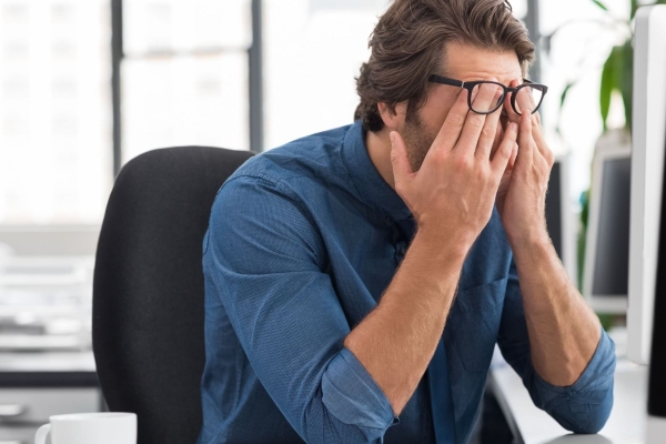 A white man with short brown hair holds his head in his hands in front of a computer monitor in a plain office. He wears a blue shirt and his glasses are pushed on to his forehead