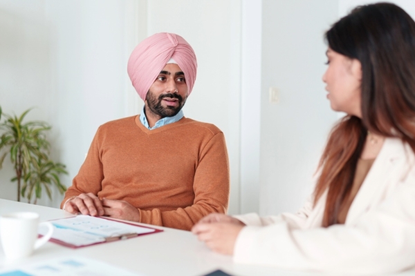 An Asian man wearing a deep-orange jumper and pink turban in conversation around a table with a woman with long dark hair and white jacket.