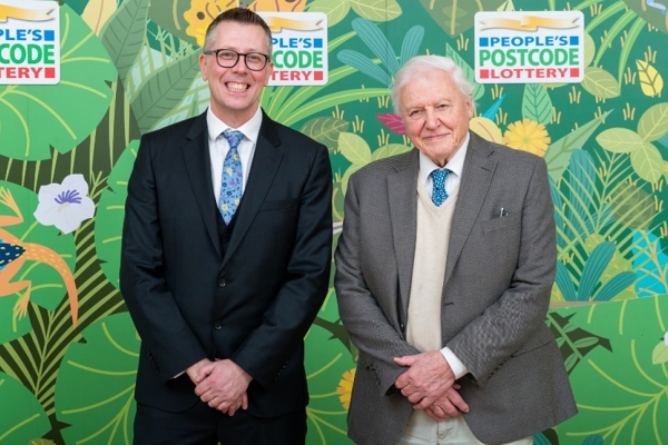 David Miller and Sir David Attenborough stood against a background composed of illustrated leaves and fauna. Both are white men with short hair wearing suits.