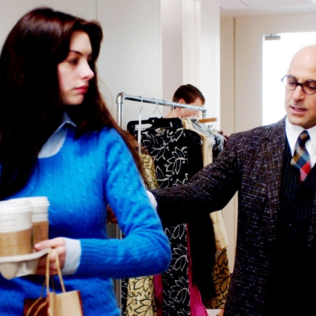 A still from the film Devil Wears Prada featuring Anne Hathaway in a blue jumper holding a tray of coffees while Stanley Tucci in a suit is stood behind her.