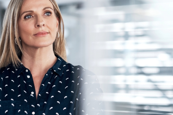 A blonde woman wearing a black blouse with white flecks looks with contemplation out of a window. The background is blurred.