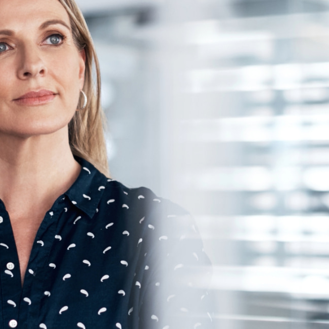 A blonde woman wearing a black blouse with white flecks looks with contemplation out of a window. The background is blurred.