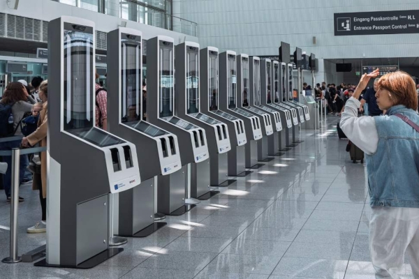 The back of a person wearing a blue top and white trousers looking at a row of tall, grey coloured computer scanners in a spacious airport.